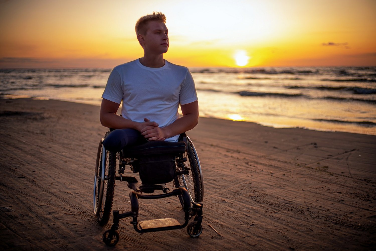 Handicapped man in wheelchair alone on a beach at sunset.
