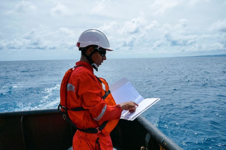 Safety officer, standing on an industrial ship, wearing overalls, a helmet, safety goggles and holding a clipboard with checklists.