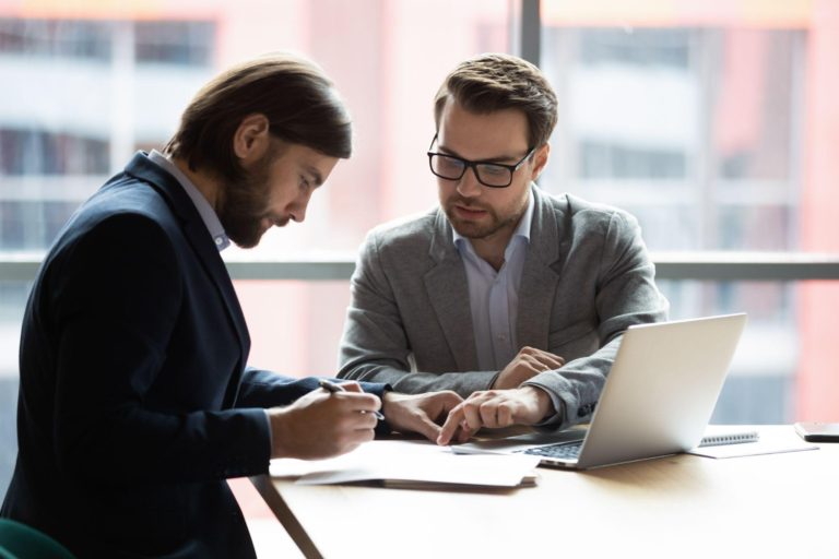 Focused young businessman signing agreement with skilled lawyer in eyeglasses.