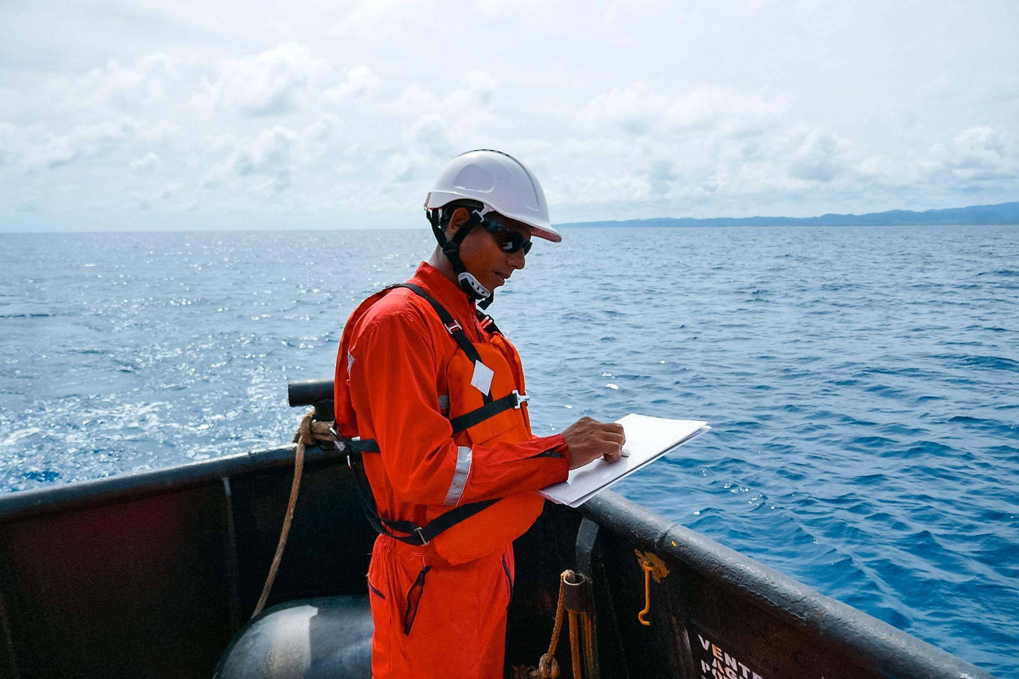 Safety officer, standing on an industrial ship, wearing overalls, a helmet, safety goggles and holding a clipboard with checklists.