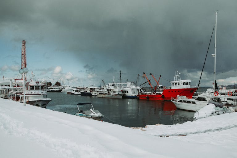 Seashore covered with snow near sea with various ships and yachts against overcast sky in port in cold winter day